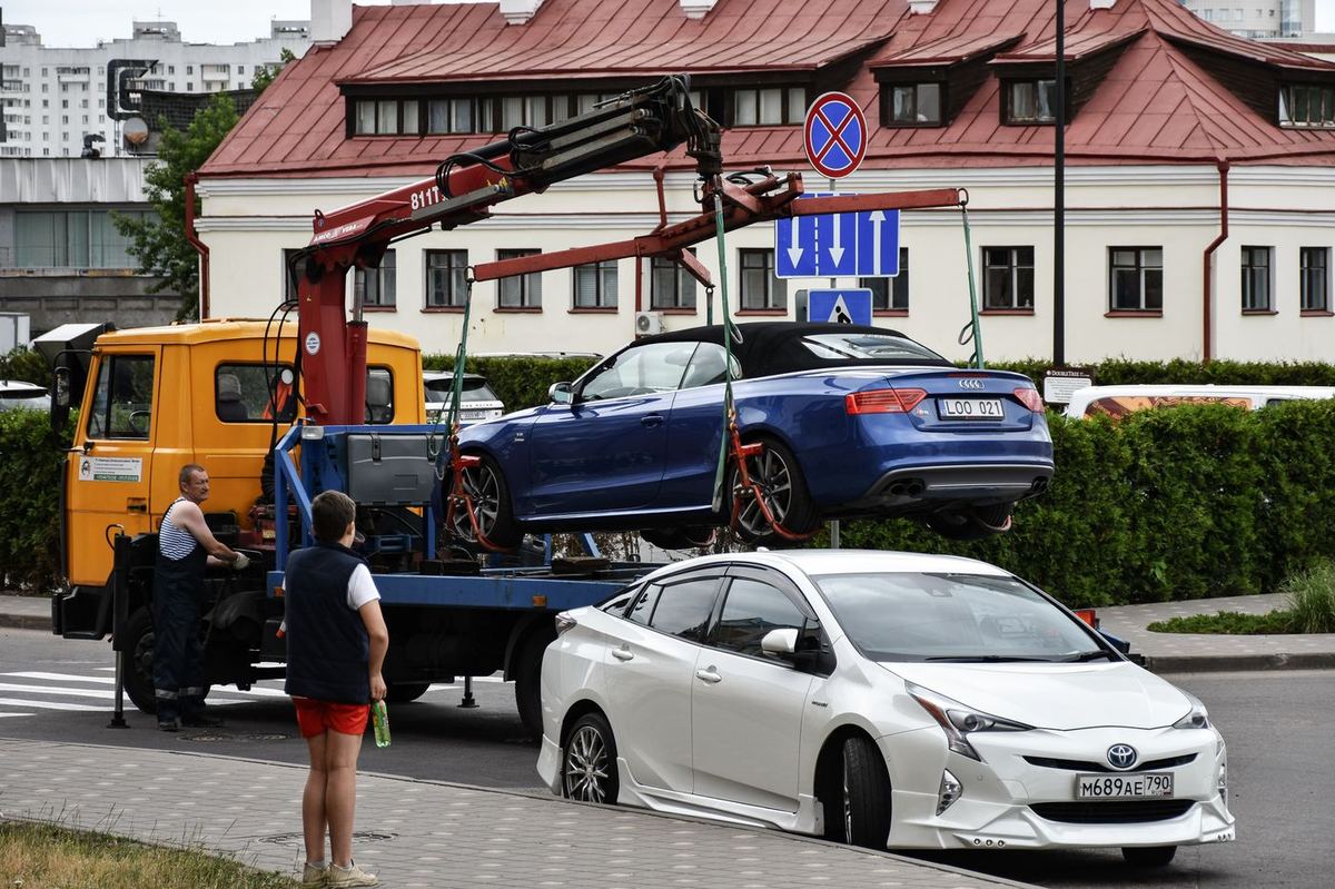 Yellow flatbed tow truck using a crane to lift a blue convertible on a city street.